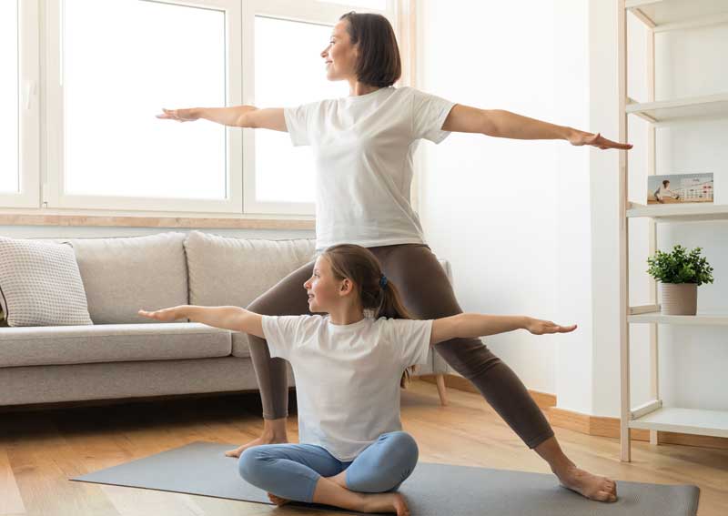A woman and a young girl practicing yoga in a living room, both performing a warrior pose on yoga mats near a sofa and a bookshelf - corrective exercises