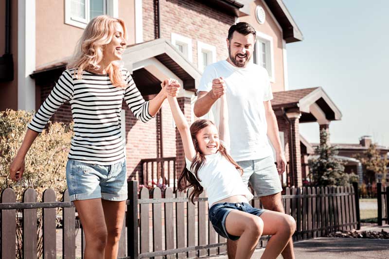 A smiling family is swinging their daughter by the arms while walking in front of houses - Chiropractor Langhorne PA A smiling family is swinging their daughter by the arms while walking in front of houses - Chiropractor Langhorne PA