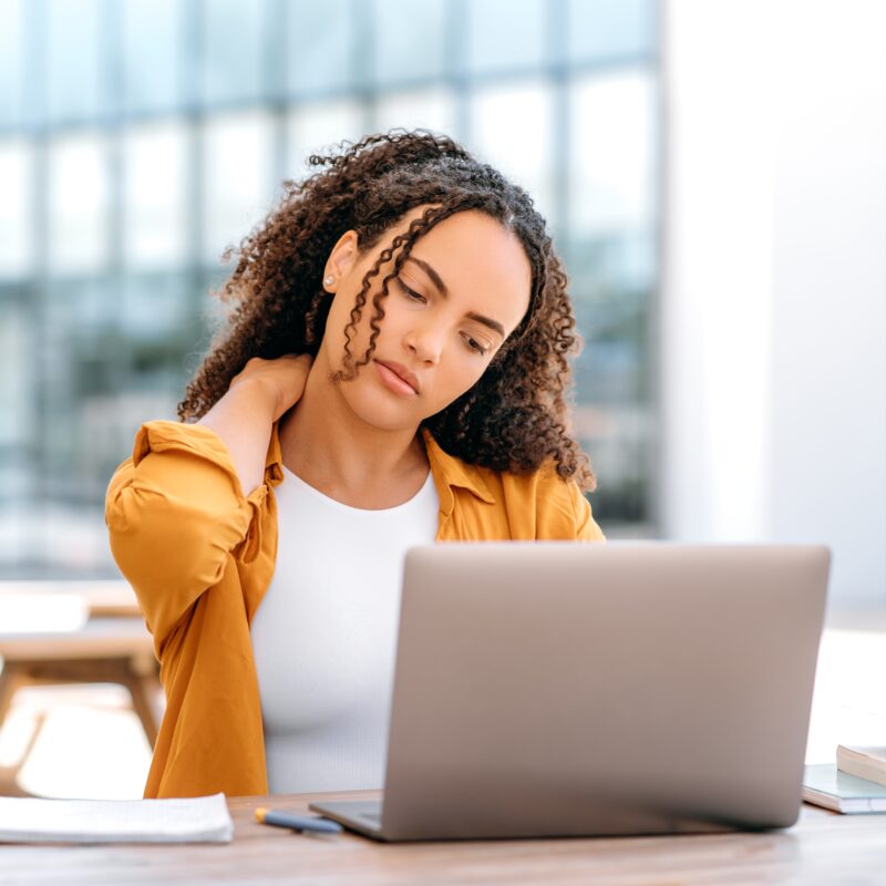 A woman seated outdoors at a table with a laptop, holding her neck in discomfort, likely from poor posture or tech strain - Neck Problems