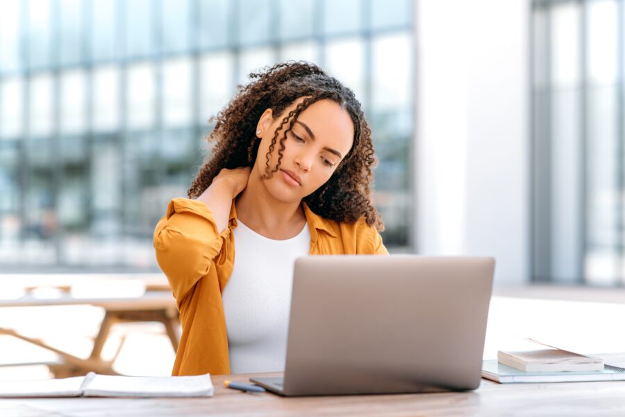 A woman seated outdoors at a table with a laptop, holding her neck in discomfort, likely from poor posture or tech strain - Neck Problems