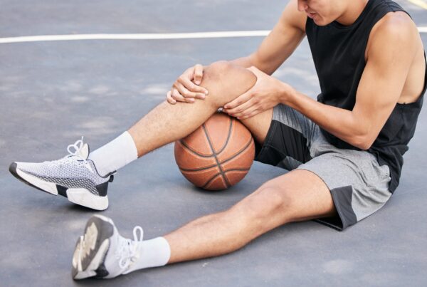 A young male athlete sitting on an outdoor basketball court, clutching his knee in pain beside a basketball, highlighting the need for rehabilitation - Spine and Sports Therapy