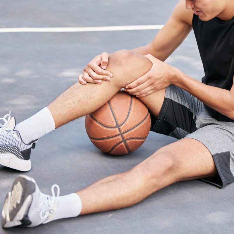 A young male athlete sitting on an outdoor basketball court, clutching his knee in pain beside a basketball, highlighting the need for rehabilitation - Spine and Sports Therapy