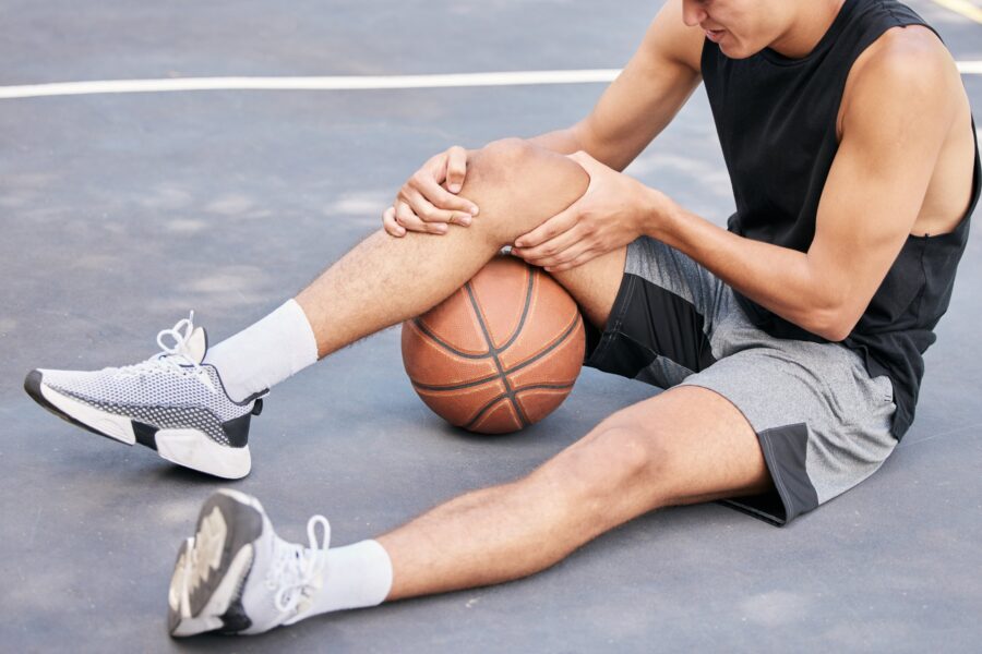 A young male athlete sitting on an outdoor basketball court, clutching his knee in pain beside a basketball, highlighting the need for rehabilitation - Spine and Sports Therapy