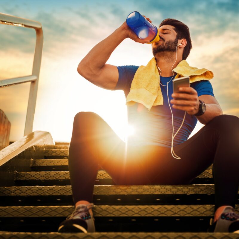 A fit man sitting on outdoor steps drinking water after a workout, holding a phone with headphones and towel, post-corrective exercise session - Corrective Exercises