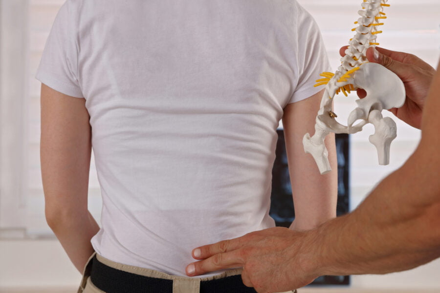 A patient stands facing away while a chiropractor points to their lower back, using a spine model to explain alignment during a spinal screening - Spinal Screening