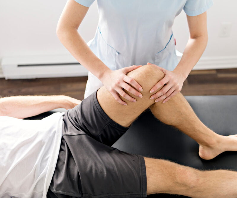A rehabilitation specialist assisting a patient with manual knee therapy on a treatment table to improve joint function and mobility - Physical Rehabilitation