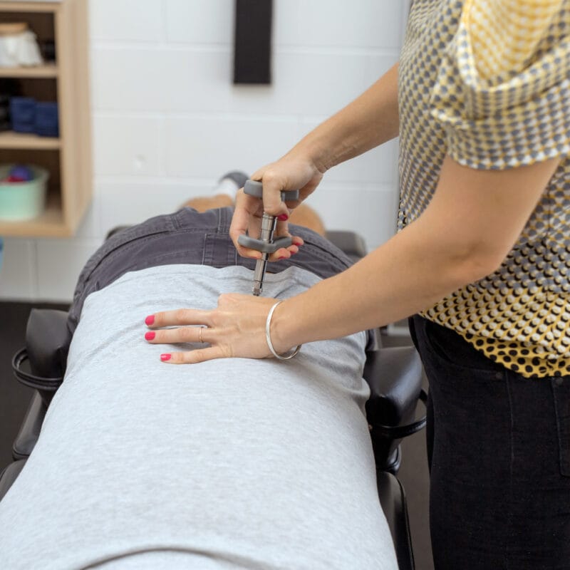 A chiropractor applying a chiropractic activator tool to a patient’s lower back while they lie face down on a treatment table - Chiropractic Activator
