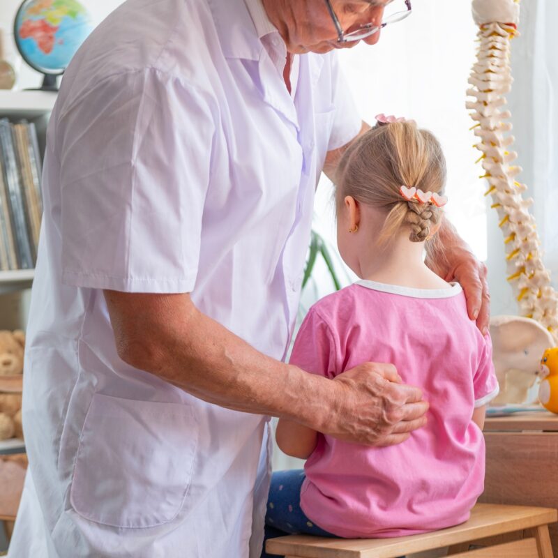 A pediatric chiropractor gently assessing a young girl’s back posture in a child-friendly clinic with anatomical models nearby - Children’s Chiropractor