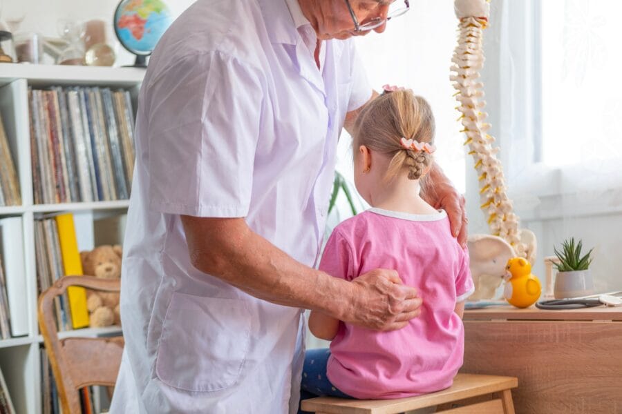 A pediatric chiropractor gently assessing a young girl’s back posture in a child-friendly clinic with anatomical models nearby - Children’s Chiropractor