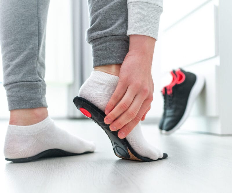 A person wearing white socks inserting a black and red orthotic insole into a running shoe, preparing for foot support - Orthotics