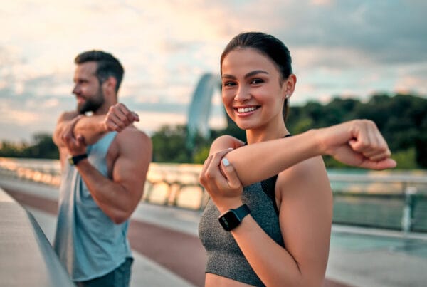 A smiling woman and man stretching their arms during an outdoor fitness session on a bridge at sunrise, demonstrating healthy posture - Corrective Exercise