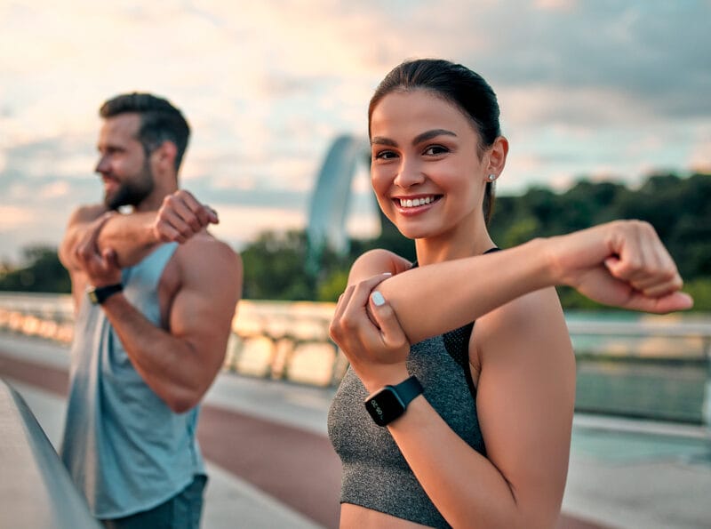A smiling woman and man stretching their arms during an outdoor fitness session on a bridge at sunrise, demonstrating healthy posture - Corrective Exercise