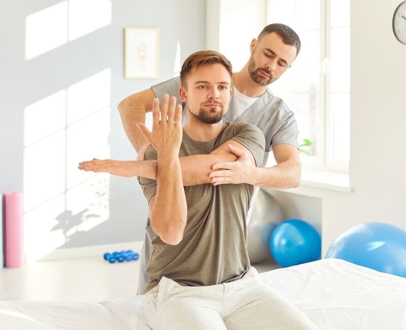 A chiropractor guiding a male patient through a shoulder mobilization technique during a therapy session in a bright clinic - Extremity Adjusting