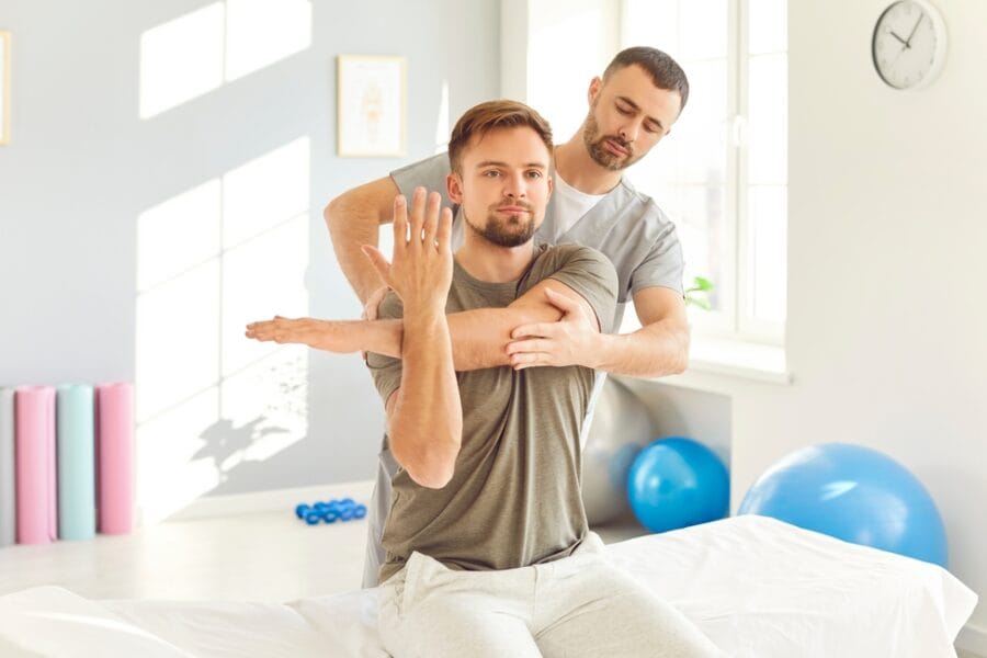 A chiropractor guiding a male patient through a shoulder mobilization technique during a therapy session in a bright clinic - Extremity Adjusting